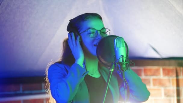 Uma jovem mulher de óculos e auscultadores a cantar no estúdio. Iluminação de néon — Vídeo de Stock