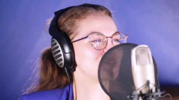 Eine junge lächelnde Frau mit Kopfhörern, die im Studio ein Lied singt — Stockvideo