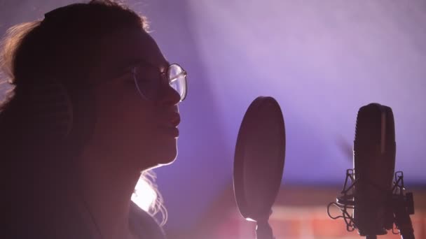 Μια νεαρή γυναίκα με γυαλιά και ακουστικά που στέκονται δίπλα στο μικρόφωνο και τραγουδούν ένα τραγούδι — Αρχείο Βίντεο