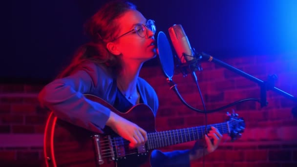 Een jonge vrouw in een bril gitaar spelen en zingen lied in neon verlichting — Stockvideo