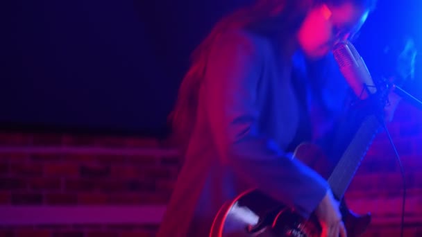 Een jonge vrouw gaat zitten en begint te spelen gitaar en zingen in neon verlichting — Stockvideo