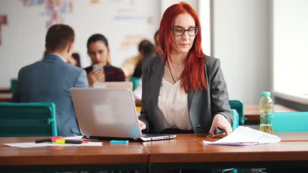 En ingefära kvinna i glasögon sitta på kontoret och arbeta av den bärbara datorn. Kontrollera hennes telefon och flytta den bort — Stockvideo