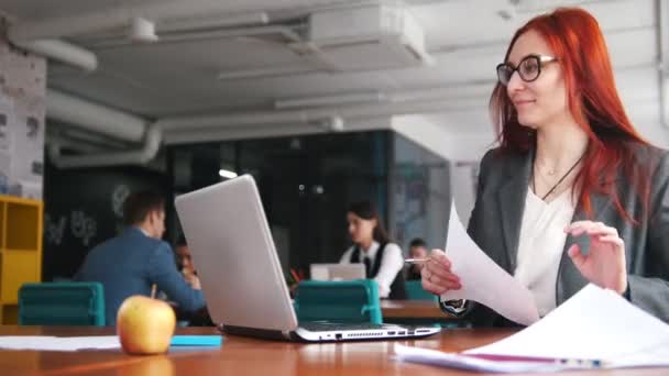 Una donna rossiccia seduta in ufficio e che lavora vicino al portatile e prende appunti — Video Stock