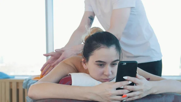 Massage session. Young woman sitting in her phone while receiving a relaxing massage