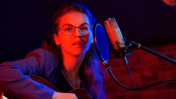 Eine junge hübsche Frau mit Brille, die Gitarre spielt und in Neonlicht singt — Stockfoto