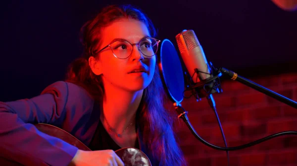 Eine junge Frau mit Brille, die Gitarre spielt und in Neonlicht singt — Stockfoto