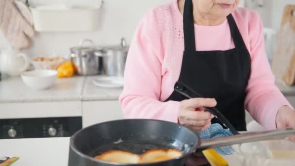 Een oude vrouw pannenkoeken maken in de lichte keuken — Stockvideo