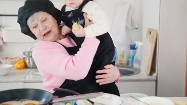 Een oude vrouw pannenkoeken maken in de keuken en een klein meisje te houden op de handen — Stockvideo