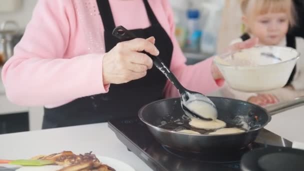 Een oude vrouw pannenkoeken maken in de keuken. Leg het deeg op de pan. Een andere oude vrouw met een klein meisje op haar handen — Stockvideo