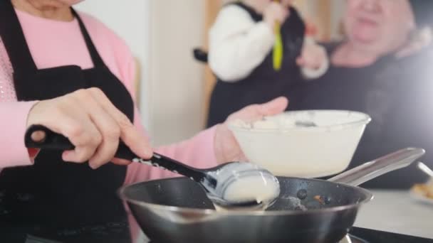 Старуха делает блинчики на яркой кухне. Положи тесто на сковородку. Еще одна пожилая женщина держит маленькую девочку на руках — стоковое видео