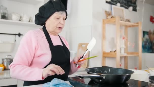 오래 된 여자는 밝은 부엌에서 팬케이크를 만들기. 접시에 팬케이크를 퍼 팅 — 비디오