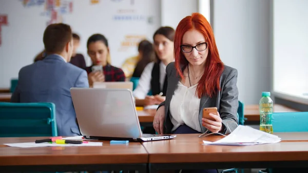 Una mujer de jengibre con gafas sentada en la oficina y trabajando junto al portátil. Comprobando su teléfono — Foto de Stock