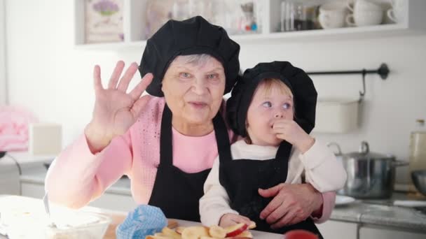 Seorang wanita tua dengan seorang gadis kecil melambaikan tangan dan melihat ke kamera di dapur . — Stok Video
