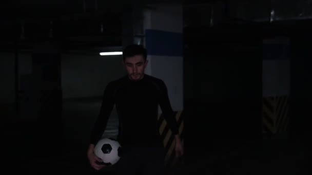 Un joven en el estacionamiento subterráneo que viene de la oscuridad sosteniendo una pelota de fútbol — Vídeo de stock