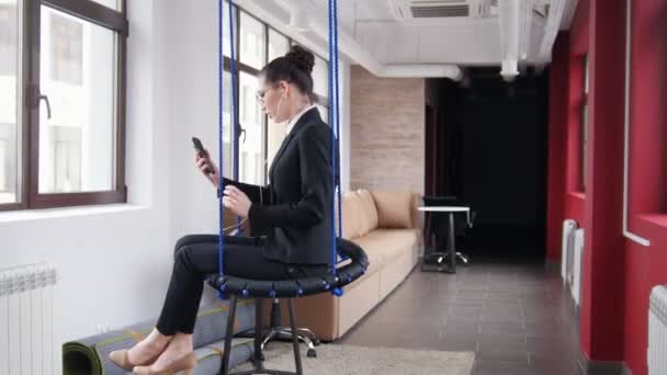 Бизнес-концепция. Женщина сидит на качелях в офисе и слушает музыку — стоковое видео