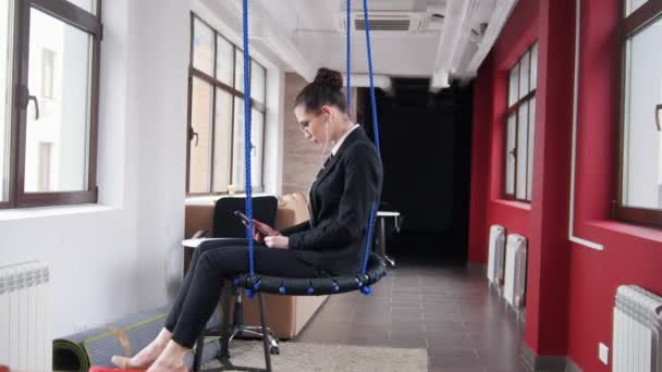 Concepto de negocio. Una mujer con gafas sentada en los columpios de la oficina y escuchando la música — Vídeos de Stock