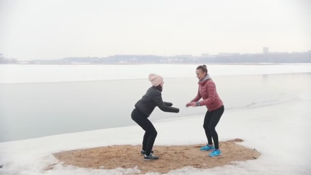 Två smala kvinnor som står på den snöiga stranden och gör knäböj — Stockvideo