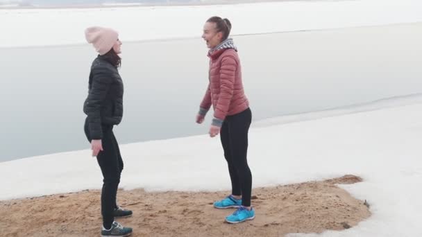 Dos mujeres delgadas en chaquetas haciendo sentadillas en la playa nevada — Vídeo de stock