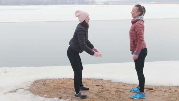 추운 날씨. 눈 덮인 해변에서 스 쿼트를 하는 재킷에 두 개의 슬림 여성 — 비디오