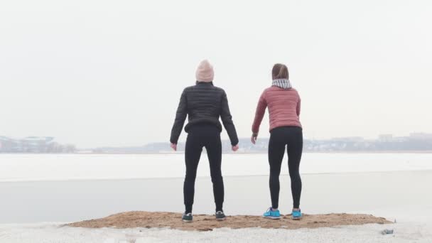 Dos mujeres delgadas en chaquetas haciendo sentadillas y inclinándose en la playa nevada — Vídeo de stock