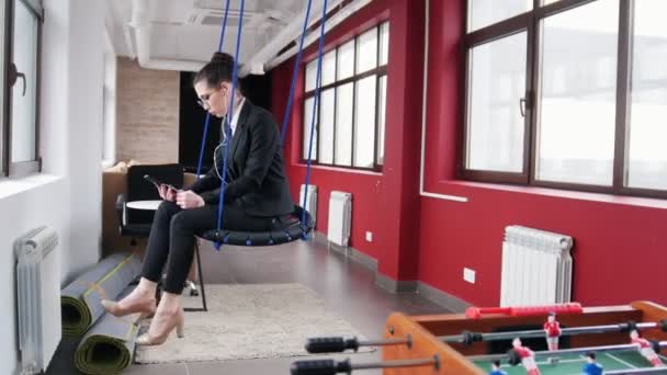 Business concept. A woman in glasses sitting on the swings in the office, listen the music and looking at the screen — ストック動画