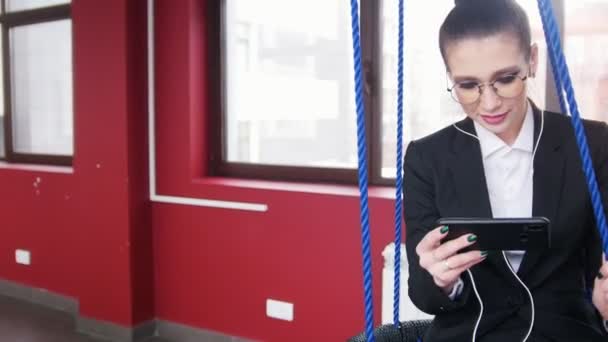 İş konsepti. Ofiste salıncaklar üzerinde oturan bir kadın, müzik dinlemek ve ekrana bakarak — Stok video