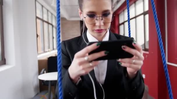 Бизнес-концепция. Молодая женщина в очках сидит на качелях в офисе, слушает музыку и смотрит на экран — стоковое видео