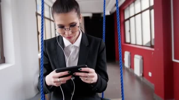 Concepto de negocio. Una mujer con gafas sentada en los columpios del centro de negocios, escuchando la música y mirando la pantalla — Vídeos de Stock