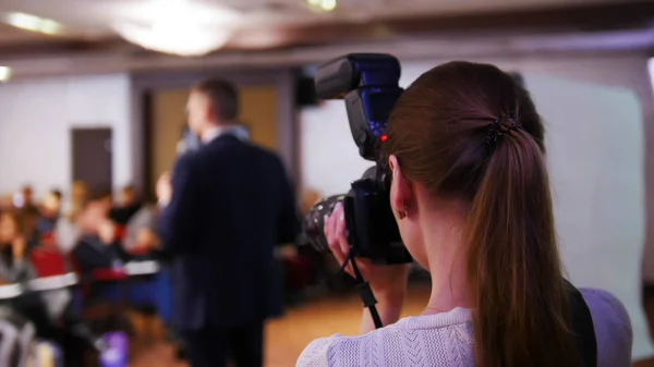 A business conference in the hall. A man talking on the stage giving a lecture. Woman with camera taking a photo of him — Stock Photo, Image