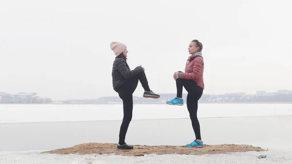 Duas mulheres magras em pé na praia nevada e se aquecendo para a corrida. Levantando as pernas — Fotografia de Stock