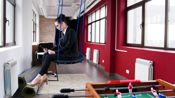 Business concept. A woman sitting on the swings in the office and listen the music — Stock Photo, Image