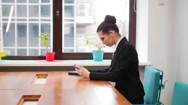 Бизнес-концепция. Женщина в очках сидит за столом и вставляет наушники в уши — стоковое фото