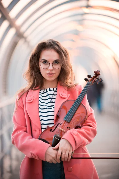 Молодая женщина в очках стоит на верхнем пешеходном переходе, держа скрипку, смотрящую в камеру — стоковое фото