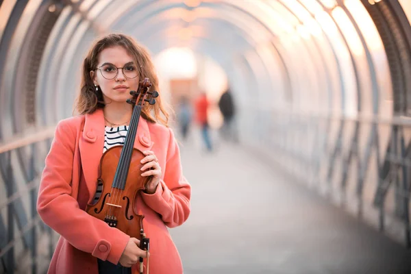 Молодая женщина в очках, стоящая на верхнем пешеходном переходе на закате, держа скрипку — стоковое фото