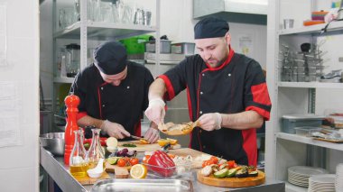 Restoran mutfağı. İki adam yemek servisi yapıyor. Bir adam balık tutuyor.