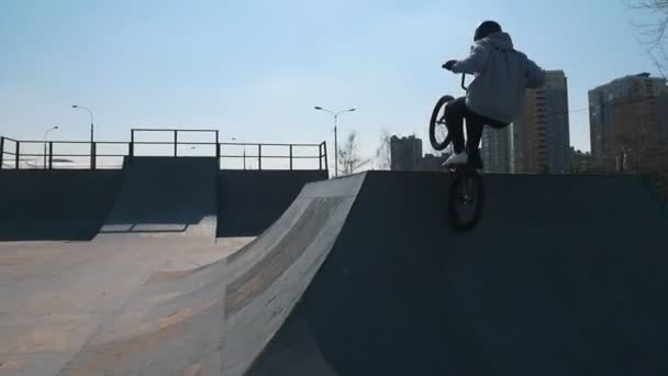 Un hombre en bicicleta haciendo trucos en el skatepark. Edificios urbanos en el fondo — Vídeos de Stock