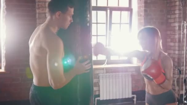 Hombre y mujer en el gimnasio. Entrenador sosteniendo un saco de boxeo y mujer golpeándolo. Un sol que sale de la ventana — Vídeo de stock