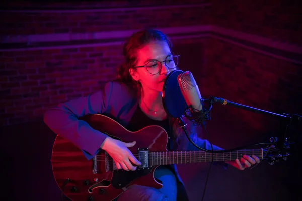 Eine junge Frau, die im Neonlicht Gitarre spielt und am Mikrofon singt — Stockfoto