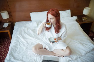 Otel odasında yatakta oturan ve çay içen zencefilli bir kadın.