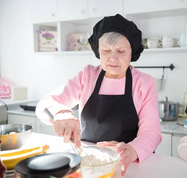 Una anciana haciendo panqueques en la cocina luminosa. Hacer masa en el tazón — Foto de Stock