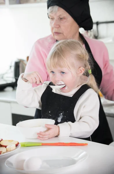 En liten flicka slarvig äta en gräddfil medan hennes farmor gör pannkakor — Stockfoto