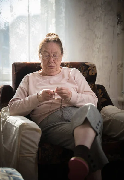 Stara kobieta w beżowym swetrze siedzącej na fotelu i dziewiarskich — Zdjęcie stockowe