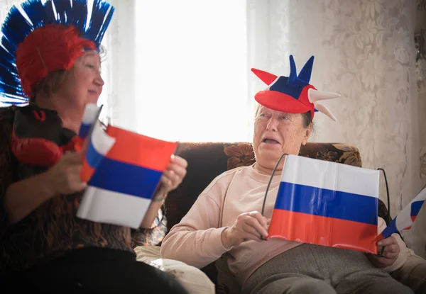 Dos ancianas entusiastas viendo televisión en accesorios rusos sosteniendo banderas rusas — Foto de Stock