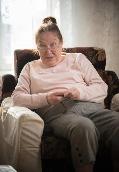 Eine alte lächelnde Frau mit Brille sitzt im Sessel und strickt. jemanden mit hochgezogenen Augenbrauen erstaunt anschauen — Stockfoto