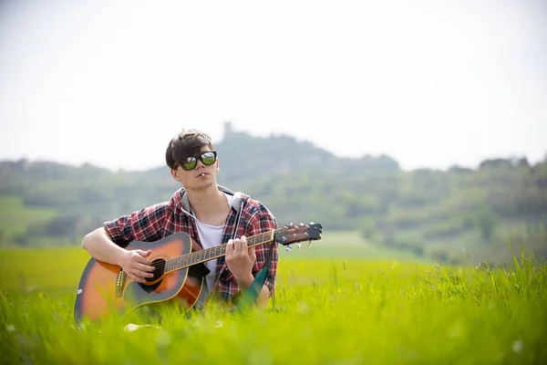 기타와 흡연을 하는 밝은 녹색 초원에 앉아 있는 선글라스를 쓴 청년 — 스톡 사진