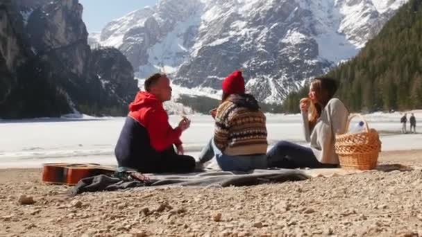 Jovens a viajar. Sentado na costa e a comer pão. Uma bela montanha no fundo — Vídeo de Stock