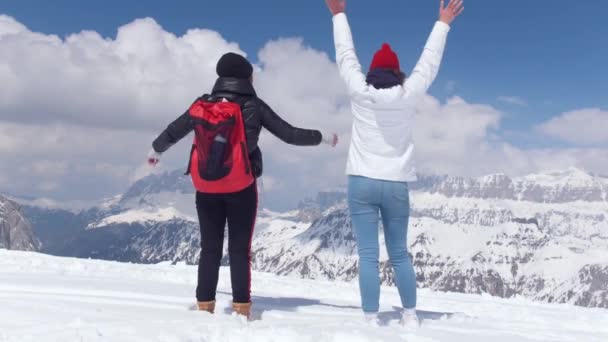 Zwei aufgeregte junge Frauen auf dem Schnee in den Dolomiten mit großem Rucksack und Gitarre und winkenden Händen — Stockvideo