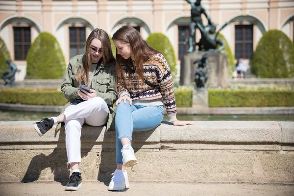 Dos mujeres jóvenes sentadas cerca de la fuente y mirando la pantalla del teléfono —  Fotos de Stock