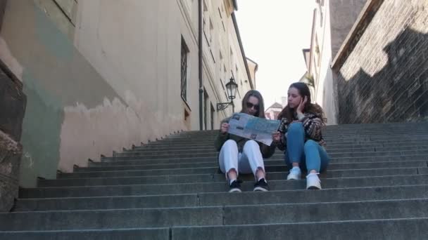 Twee jonge vrouwen zitten op de trap en kijken naar de kaart. Praten en bespreken waar te gaan volgende — Stockvideo