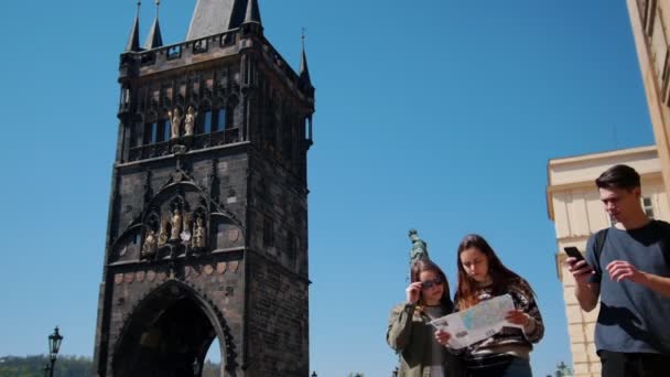 Юні друзі-мандрівники ходять на фоні вежі Карлового мосту. Жінки тримають карту і знайти шлях, щоб піти і їх друг з фотографією зору — стокове відео